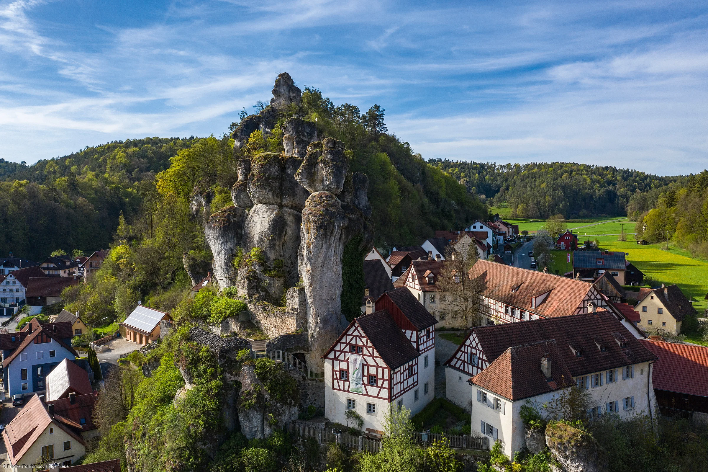 Felsenformation mit Fachwerkhäusern und Wald im Hintergrund unter blauem Himmel mit Wolken.