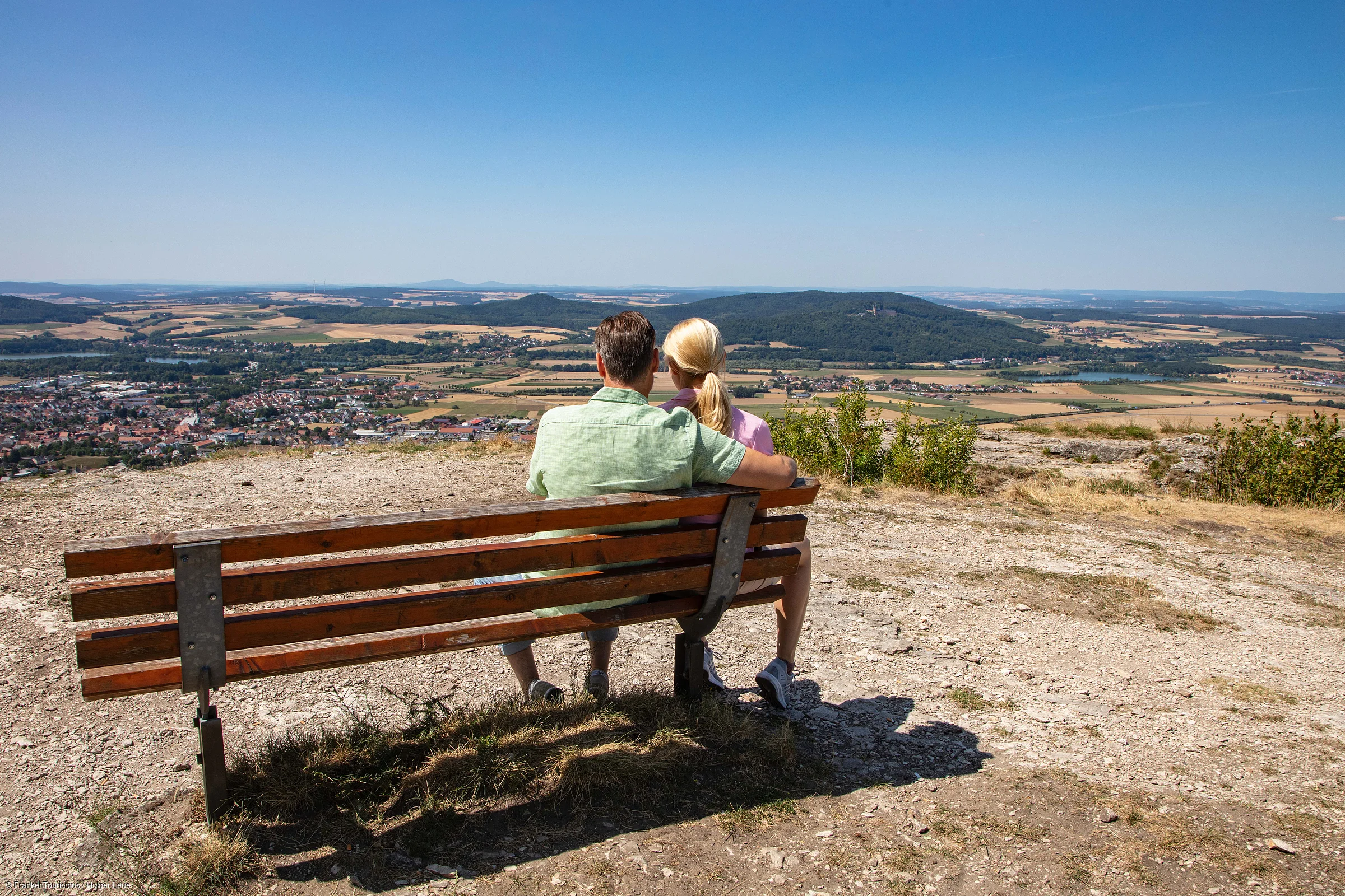 Paar sitzt auf einer Bank auf Hügel mit Blick auf Tal, Felder und Dorf unter blauem Himmel.