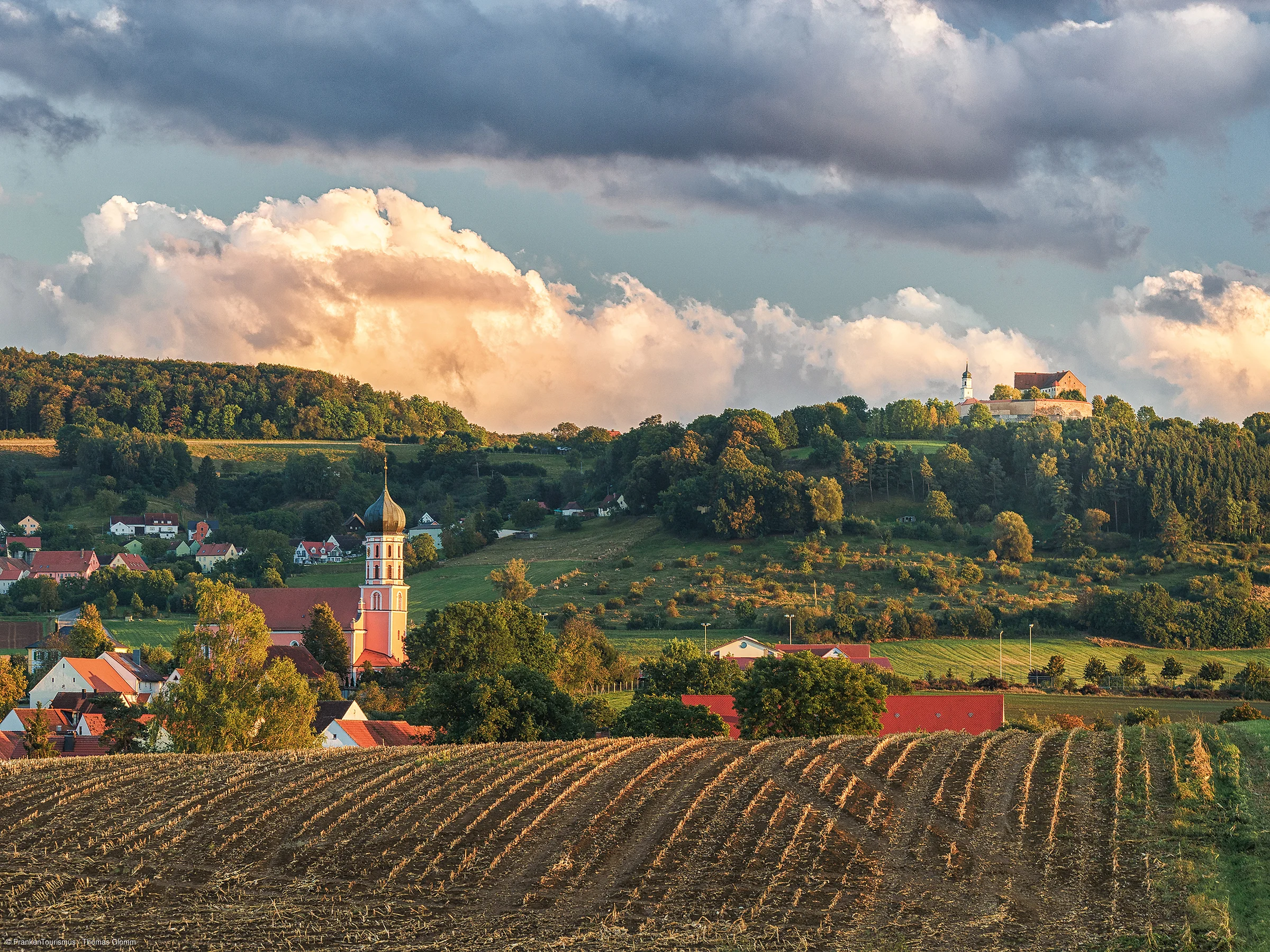 Landschaft mit abgeerntetem Feld, Dorf mit Kirche und bewaldeten Hügeln unter bewölktem Himmel.