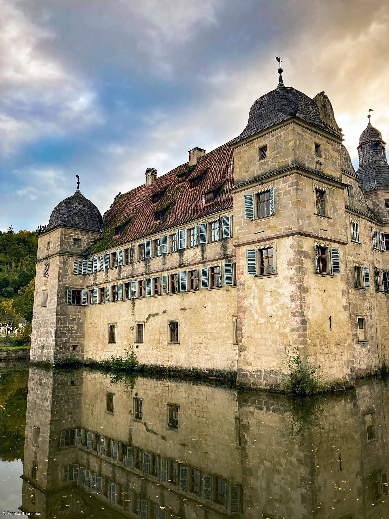 Historisches Schloss mit Türmen und blauen Fensterläden, Spiegelung im Wasser vor bewölktem Himmel