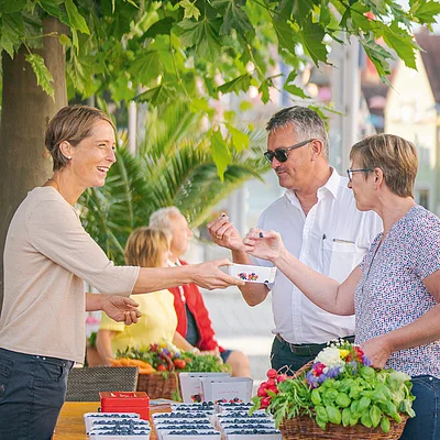 Frau verkauft Heidelbeeren an Mann und Frau mit Korb voller Gemüse an Marktstand im Freien.
