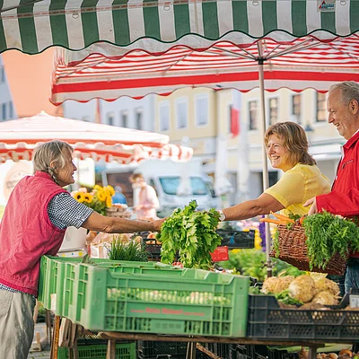 Marktstand mit Verkäuferin und zwei Kunden, die frisches Gemüse unter Sonnenschirmen tauschen.