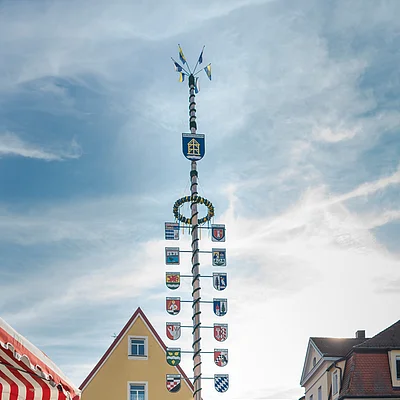 Marktplatz mit Maibaum, Blumenständen und Menschen vor gelbem Giebelhaus unter bewölktem Himmel
