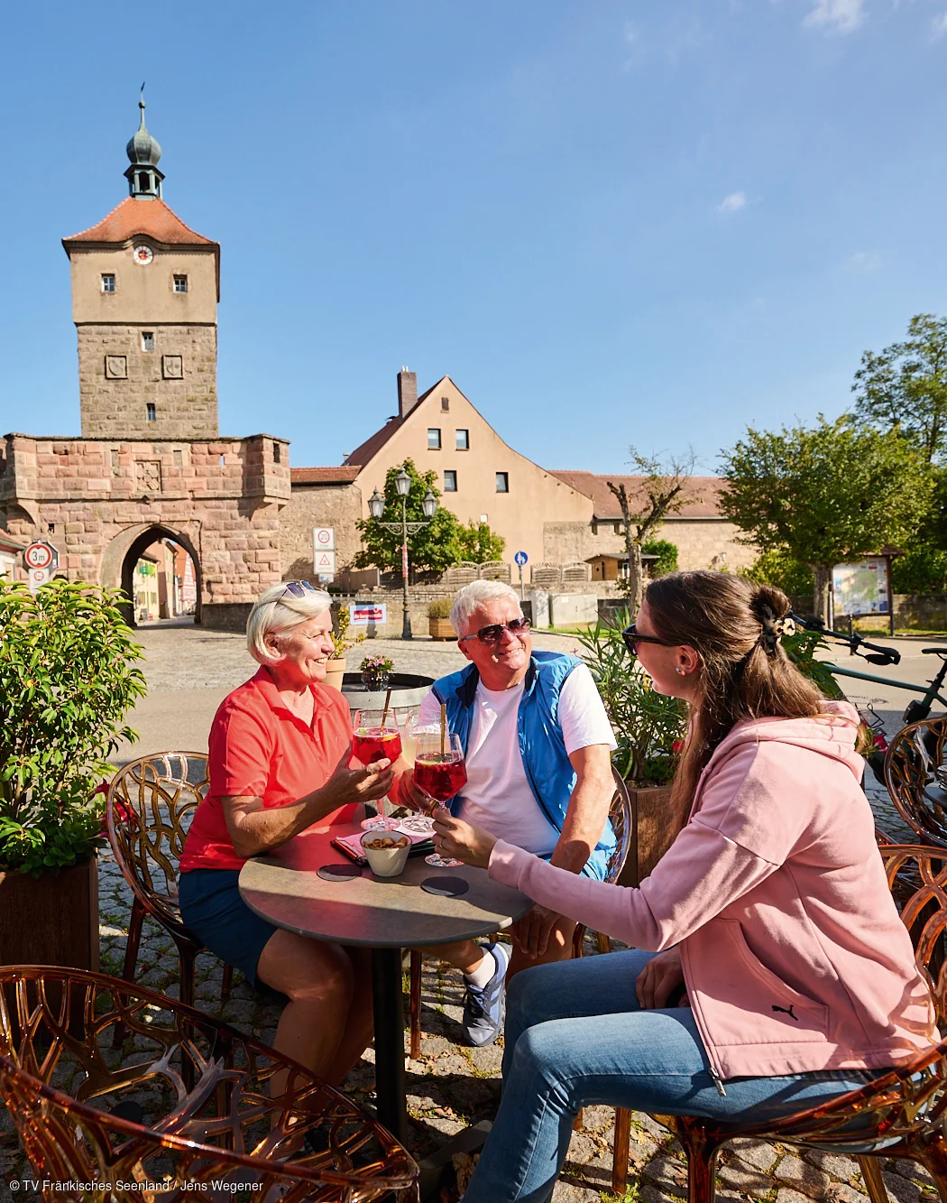 Drei Personen sitzen draußen an einem Tisch vor historischem Stadttor und stoßen mit Getränken an.
