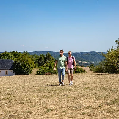 Paar hält Händchen beim Wandern auf trockenem Feld mit Kirche und Hügeln im Hintergrund.