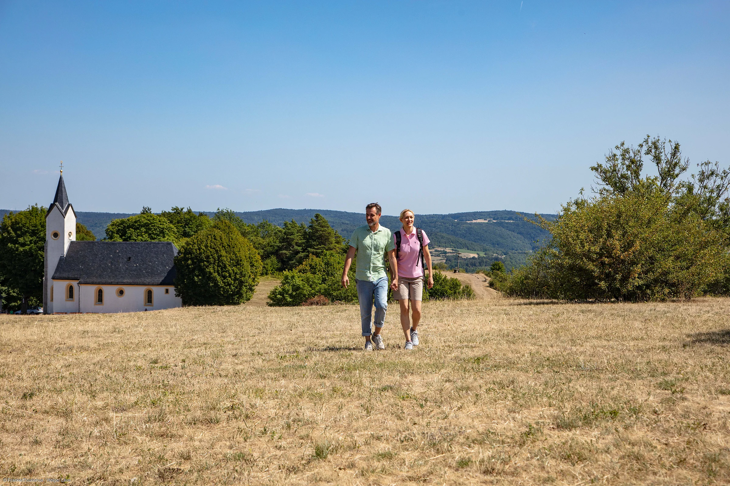 Paar hält Händchen beim Wandern auf trockenem Feld mit Kirche und Hügeln im Hintergrund.