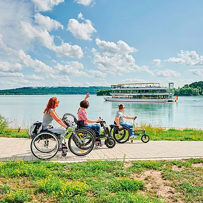 Drei Personen im Rollstuhl fahren auf einem Weg am Seeufer bei sonnigem Himmel, im Hintergrund ein Ausflugsschiff auf dem Wasser.