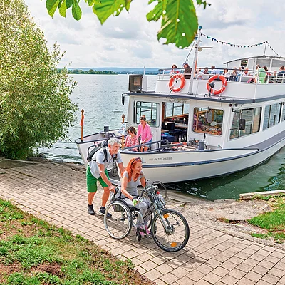 Frau im Rollstuhl mit Handbike wird von Mann geschoben, im Hintergrund ein Ausflugsboot auf dem Wasser.
