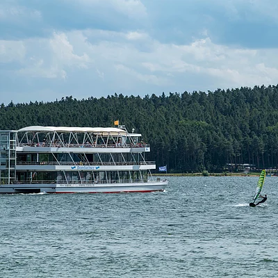 Fahrgastschiff auf See mit Windsegler und Wald im Hintergrund unter bewölktem Himmel.