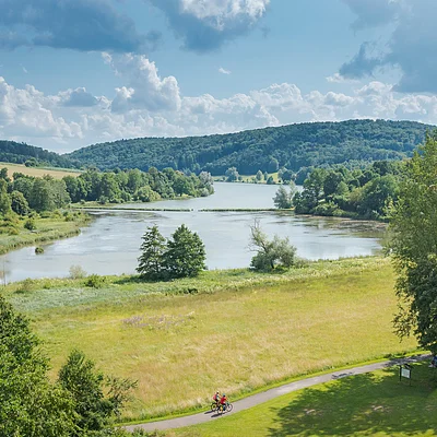 Landschaft mit See, Wiesen, Wald und zwei Radfahrern auf einem Weg im Vordergrund bei bewölktem Himmel.