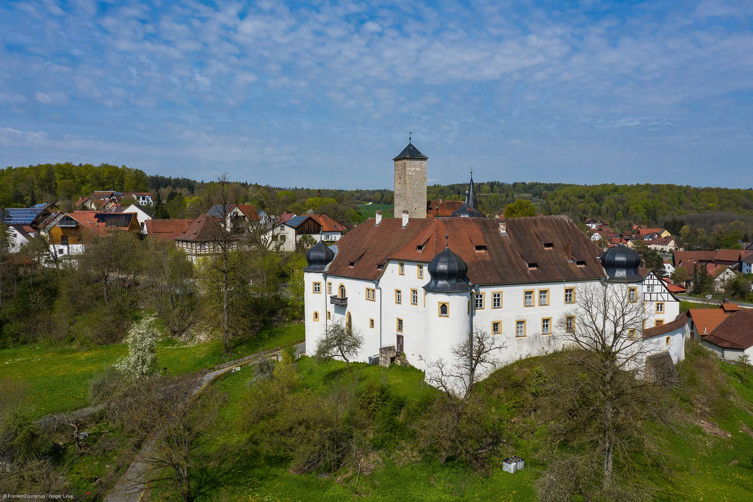 Weißes Schloss mit Türmen auf einem Hügel vor Dorf und Wald unter blauem Himmel mit Wolken