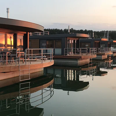 Vier moderne Hausboote mit Terrassen und Metallgeländern spiegeln sich im ruhigen Wasser bei Sonnenuntergang.