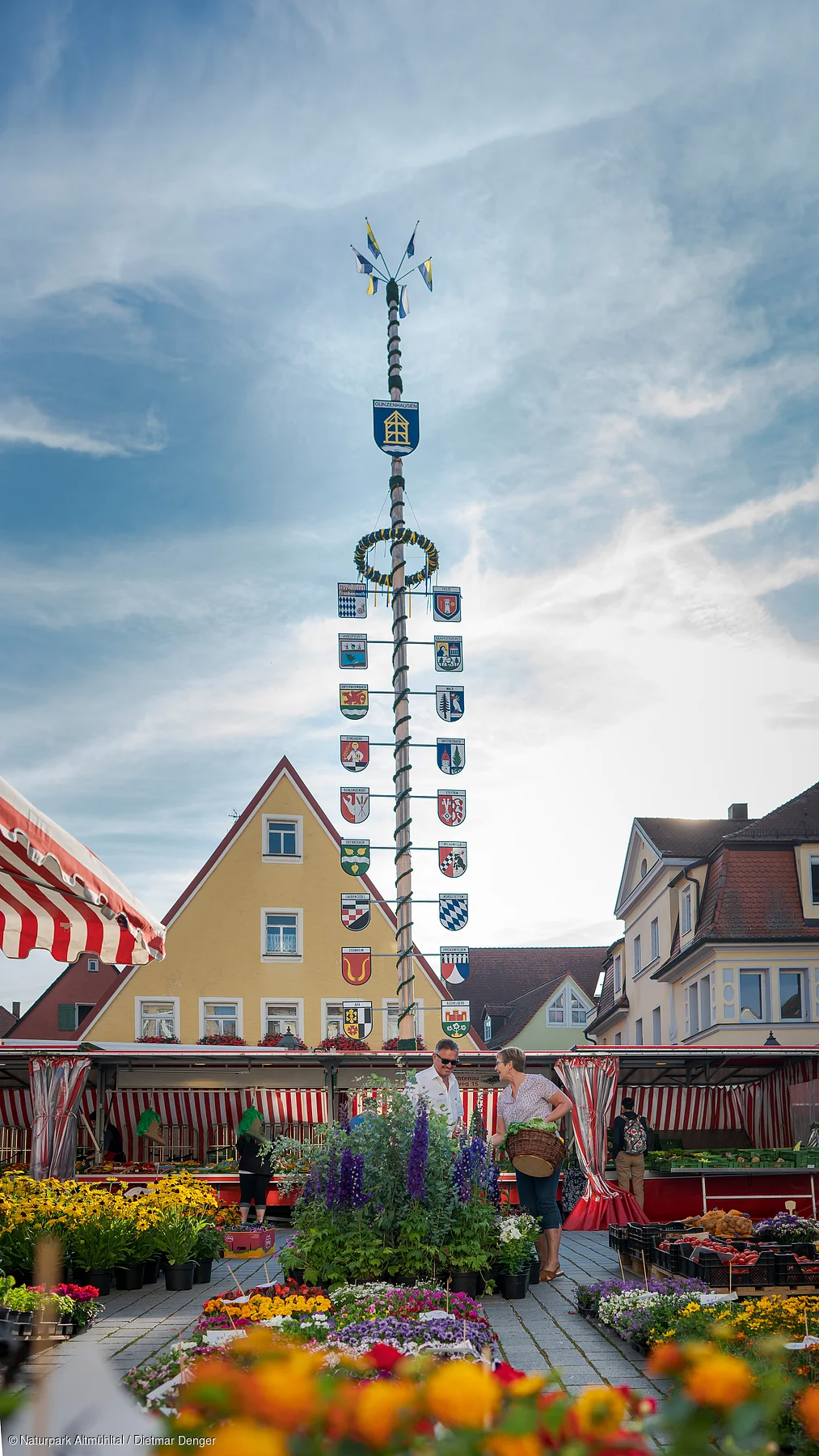 Marktplatz mit Maibaum, Blumenständen und Menschen vor gelbem Giebelhaus unter bewölktem Himmel