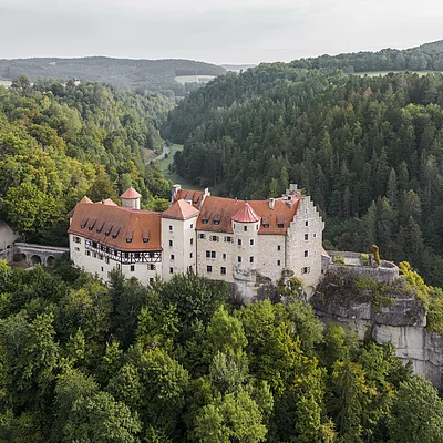 Burg mit roten Dächern auf Felsen umgeben von dichtem grünen Wald und Hügeln im Hintergrund.