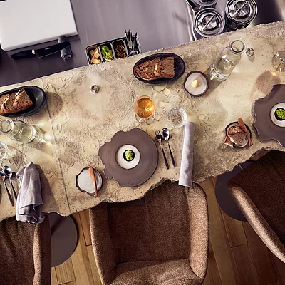 Drei gedeckte Plätze mit braunen Stühlen an einem rustikalen Steintisch mit Brot, Gläsern und Besteck.