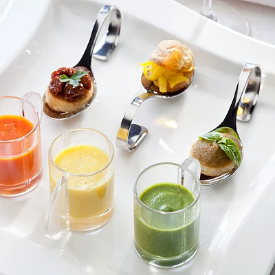 Drei kleine Gläser mit roten, gelben und grünen Saucen und drei Löffel mit kleinen Häppchen auf weißem Teller.