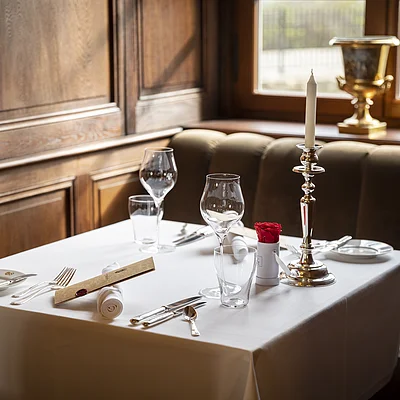 Tisch mit weißer Tischdecke, Besteck, Gläsern, Kerzenhalter mit Kerze und roter Rose vor Holzpaneelen und Fenster.