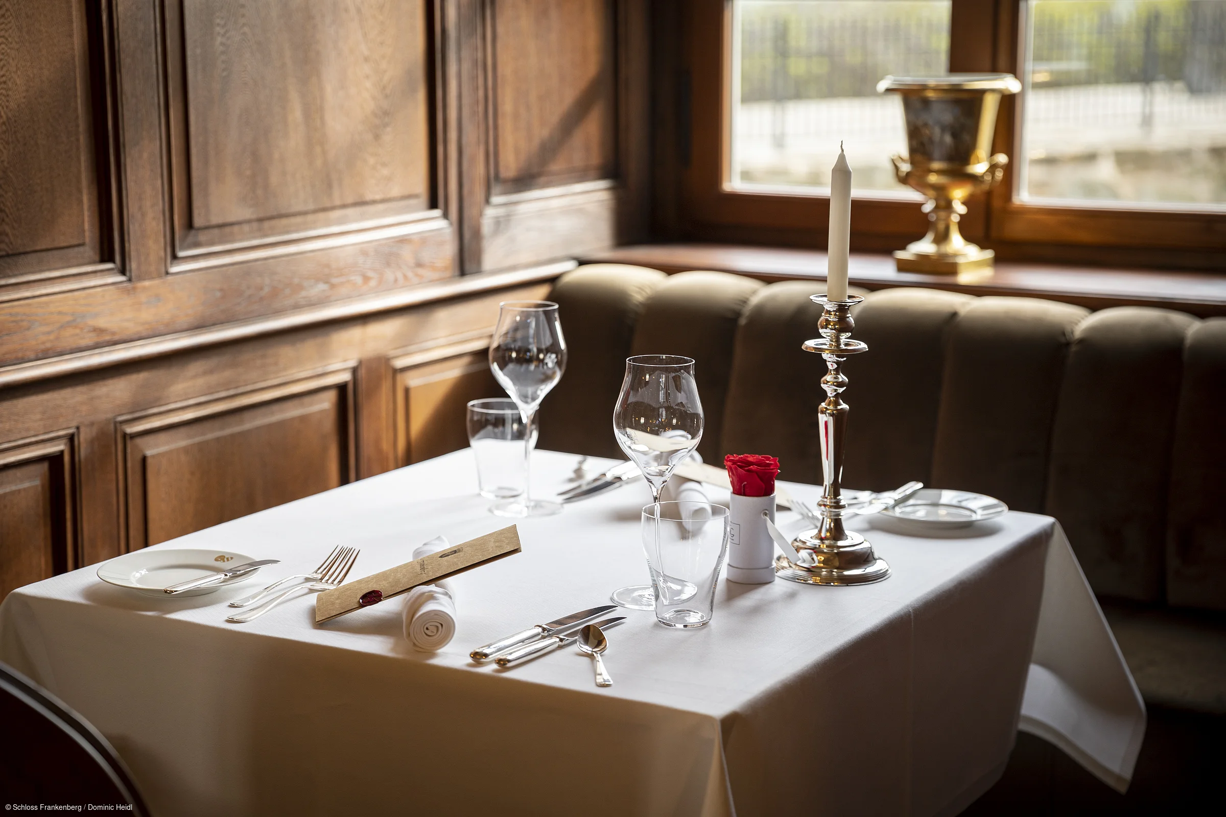 Tisch mit weißer Tischdecke, Besteck, Gläsern, Kerzenhalter mit Kerze und roter Rose vor Holzpaneelen und Fenster.