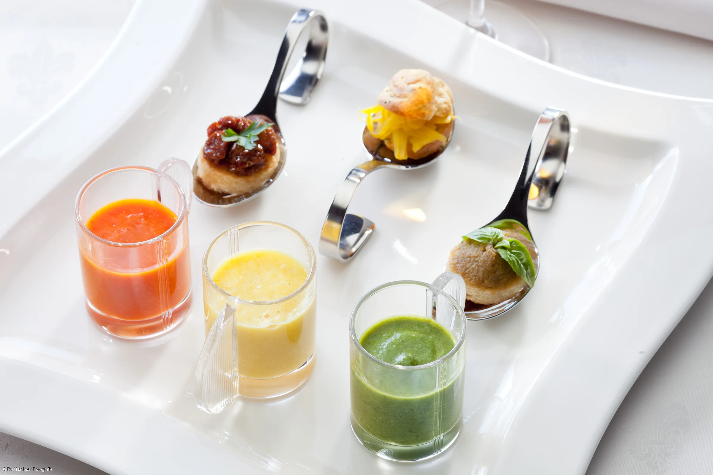 Drei kleine Gläser mit roten, gelben und grünen Saucen und drei Löffel mit kleinen Häppchen auf weißem Teller.