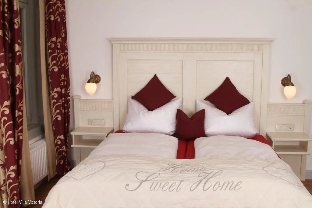 Zweibettzimmer mit weißen Bettdecken, roten Kissen, Nachttischen und roten Vorhängen mit Muster.