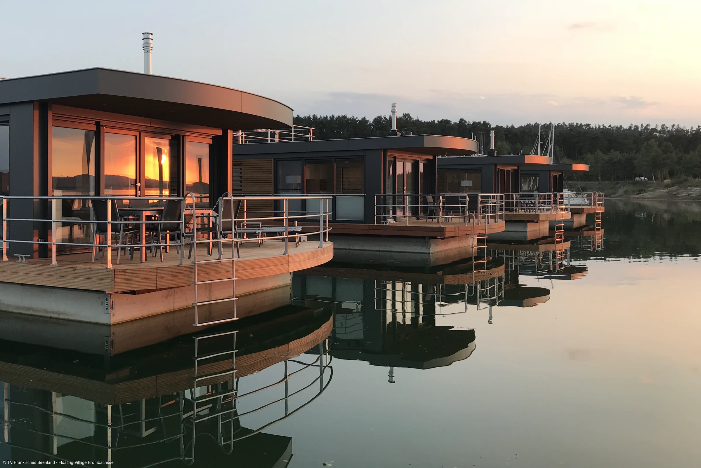 Vier moderne Hausboote mit Terrassen und Metallgeländern spiegeln sich im ruhigen Wasser bei Sonnenuntergang.