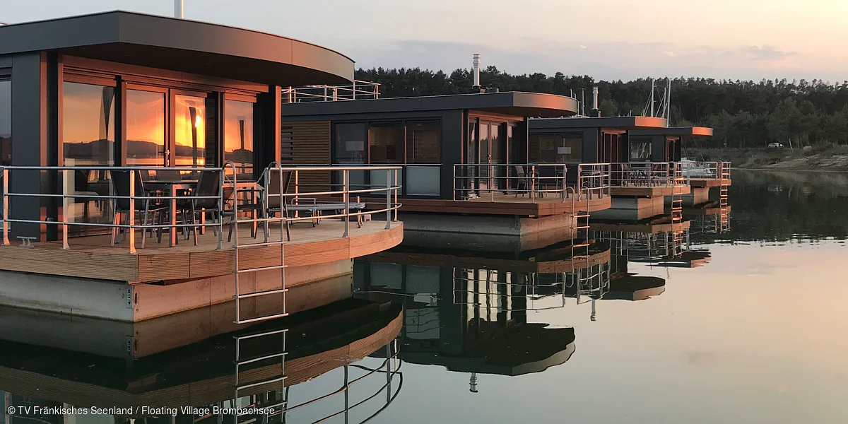 Vier moderne Hausboote mit Terrassen und Metallgeländern spiegeln sich im ruhigen Wasser bei Sonnenuntergang.
