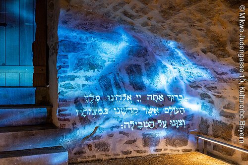 Hebräischer Text in blauer Lichtprojektion an einer Steinwand neben einer Treppe und einer Holztür