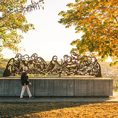 Skulptur der letzten Abendmahl-Szene auf Betonpodest im Park mit herbstlichen Bäumen und Person im Vordergrund.