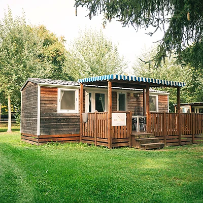 Holzhaus mit Veranda und blau-weiß gestreiftem Sonnenschutz auf grünem Rasen, umgeben von Bäumen.