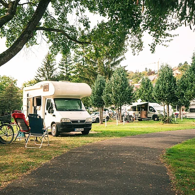 Campingplatz mit Wohnmobilen, Fahrrädern und Campingstühlen unter Bäumen neben einem Weg
