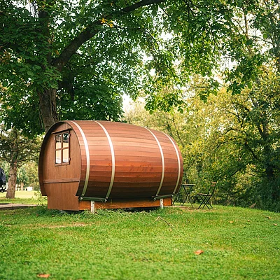 Holzhaus in Fassform auf einer Wiese unter Bäumen mit rotem Van im Hintergrund und Gartenstuhl mit Tisch