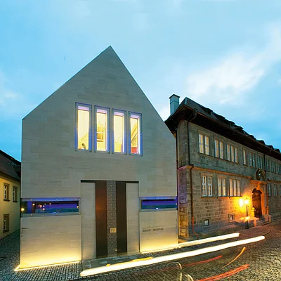 Abendaufnahme des modernen Knauf-Museums neben einem historischen Gebäude in einer gepflasterten Straße.