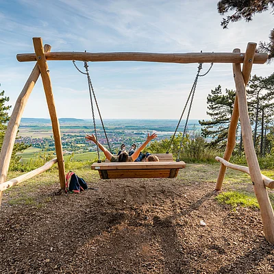 Zwei Personen sitzen auf einer großen Holzschaukel mit Blick auf ein Tal und eine Stadt im Hintergrund.