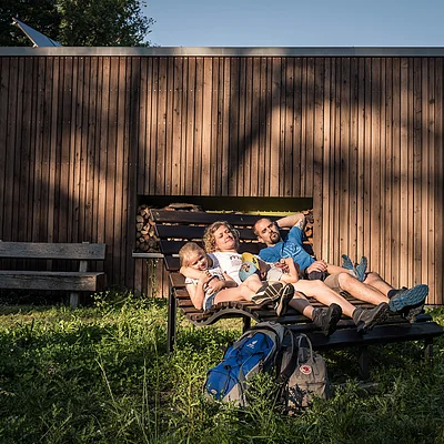 Familie mit zwei Kindern liegt auf einer Holzliege vor einem Holzhaus im Sonnenschein.
