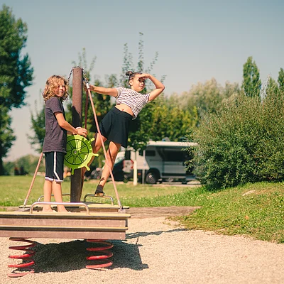 Zwei Kinder spielen auf einem Spielgerät mit Federn im Freien bei sonnigem Wetter.