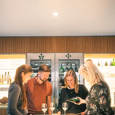 Vier Personen stehen an einem beleuchteten Tresen, eine gießt Wein in Gläser.
