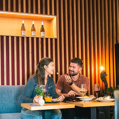 Zwei Personen sitzen an Tischen in einem Restaurant mit Holzpaneelwand und essen gemeinsam.
