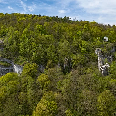 Wald mit Felsen und einem kleinen Pavillon auf einem Felsen unter blauem Himmel.