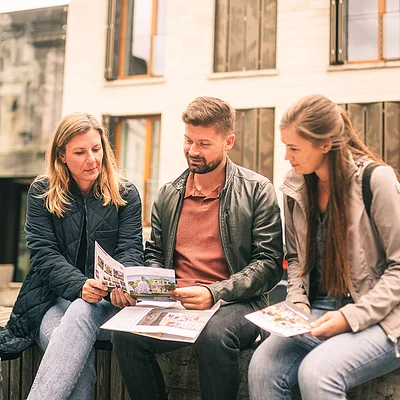 Drei Personen sitzen draußen auf einer Bank und schauen gemeinsam in Broschüren.