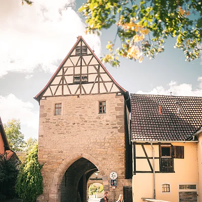 Zwei Personen gehen auf Kopfsteinpflaster vor einem historischen Torbogen mit Fachwerkhaus bei sonnigem Himmel.