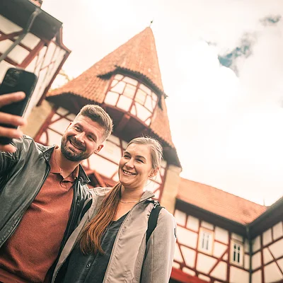 Zwei Personen machen vor einem Fachwerkhaus mit Turm ein Selfie mit dem Smartphone.