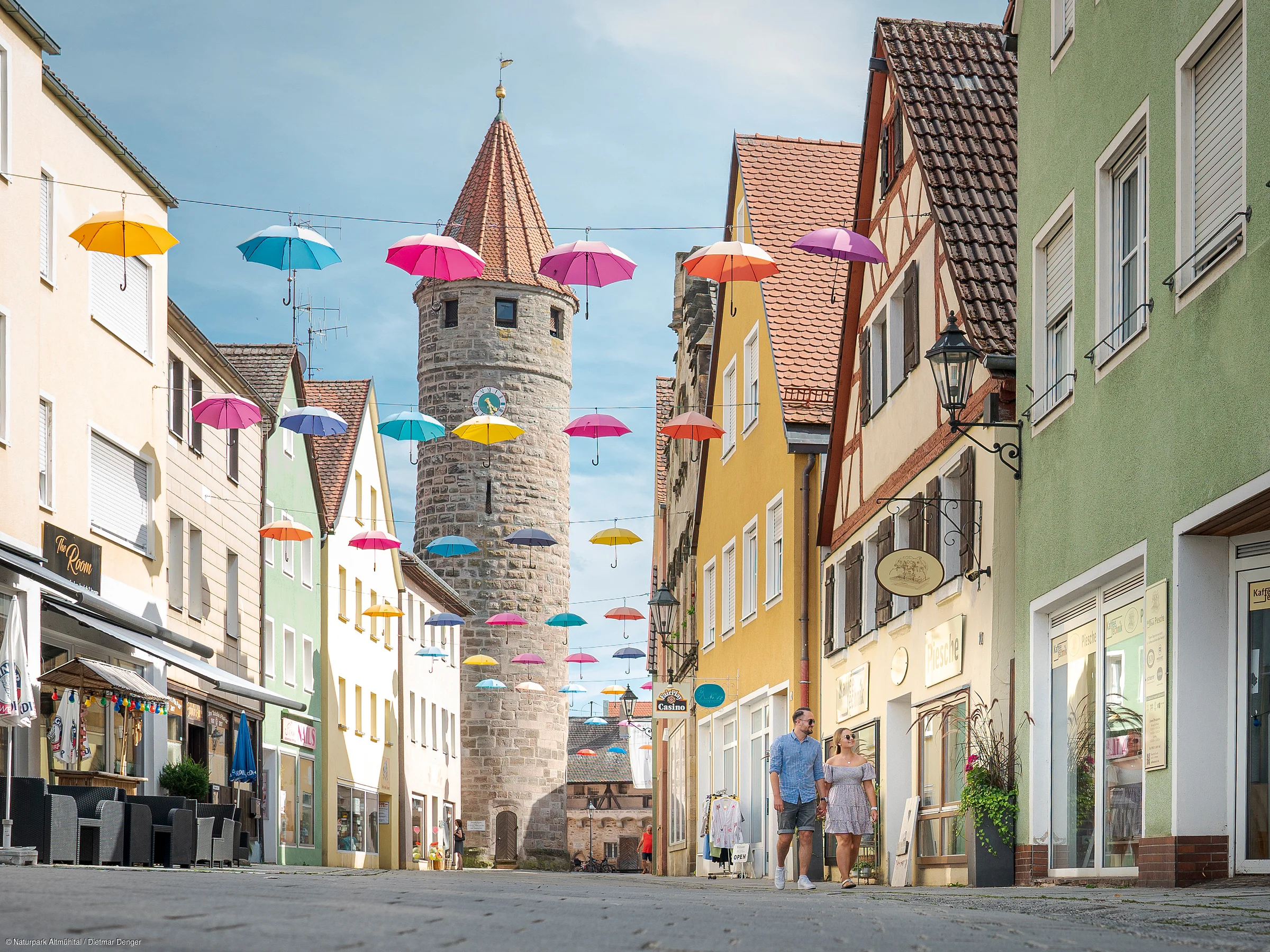 Straße mit bunten Regenschirmen über Kopf und historischem Steinturm im Hintergrund, zwei Personen gehen.