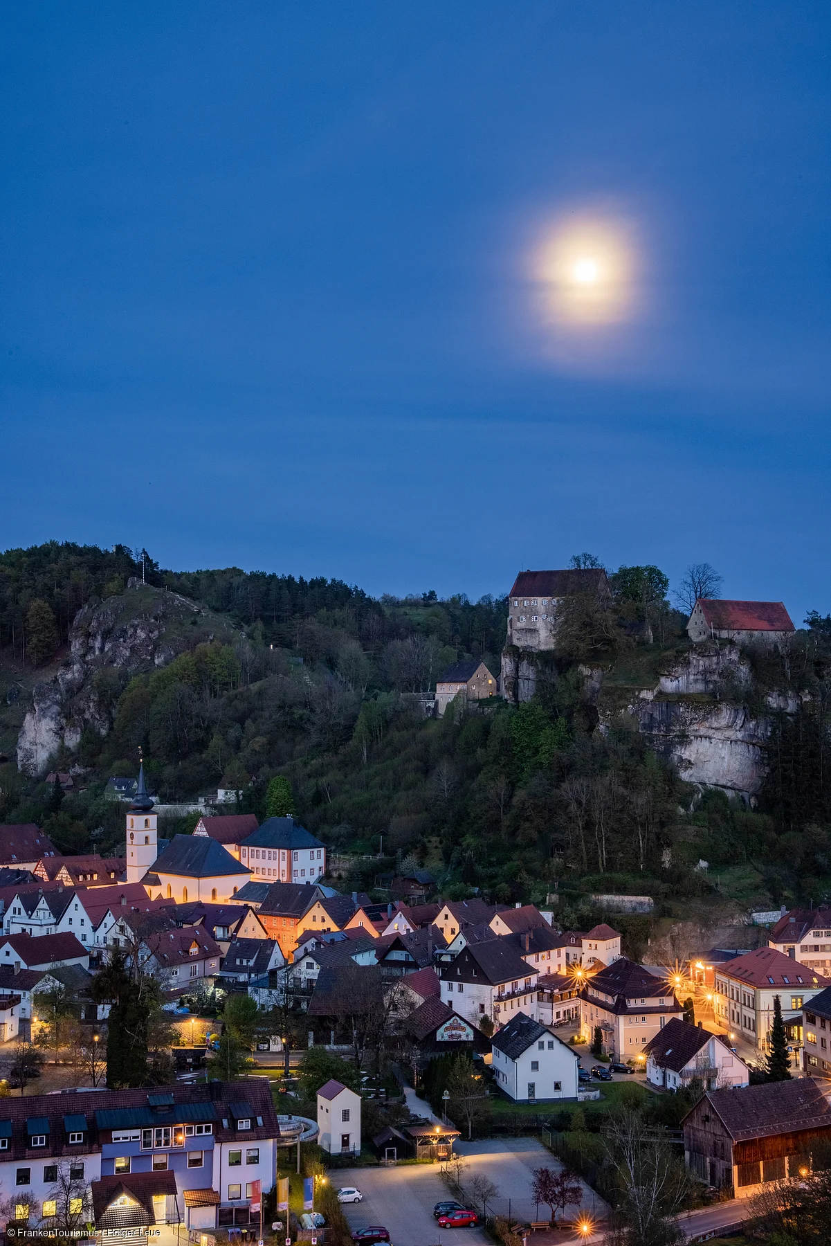 Dorf bei Nacht mit beleuchteten Häusern, Hügeln und Vollmond am klaren Himmel