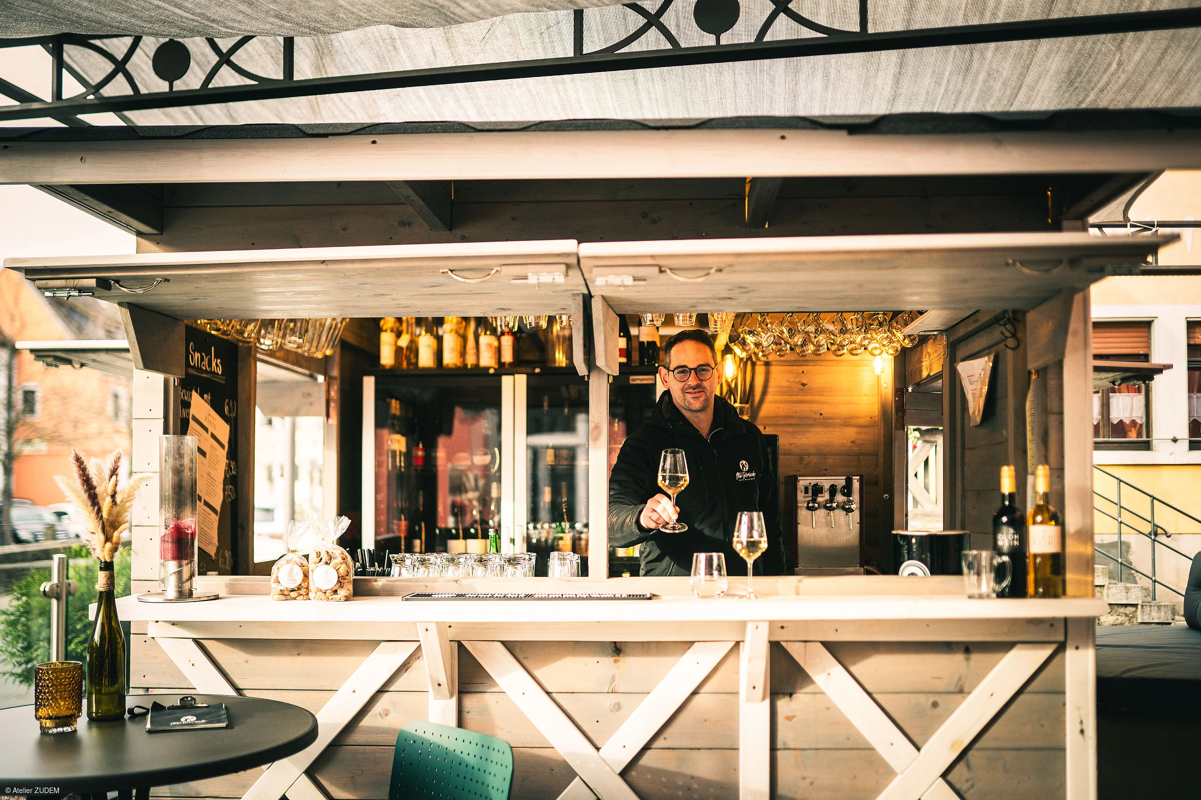 Mann mit Brille reicht Weinglas an Holzbar mit Getränken und Snacks in Außenbereich eines Lokals.