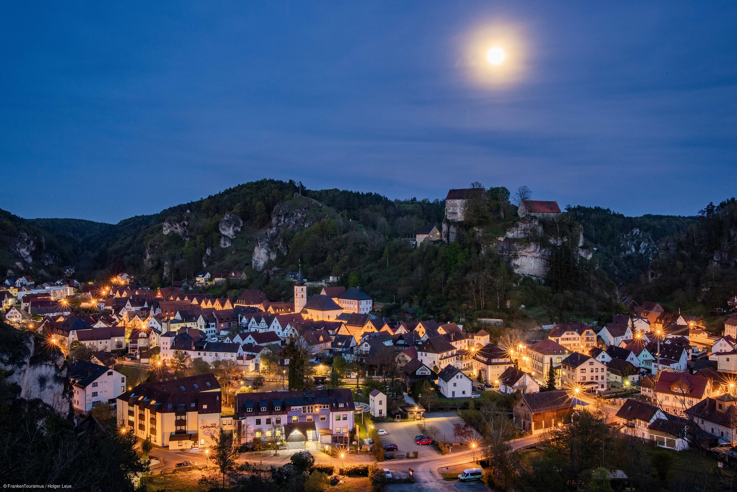 Nächtliche Stadtansicht mit beleuchteten Häusern, Hügeln und Vollmond am klaren Himmel