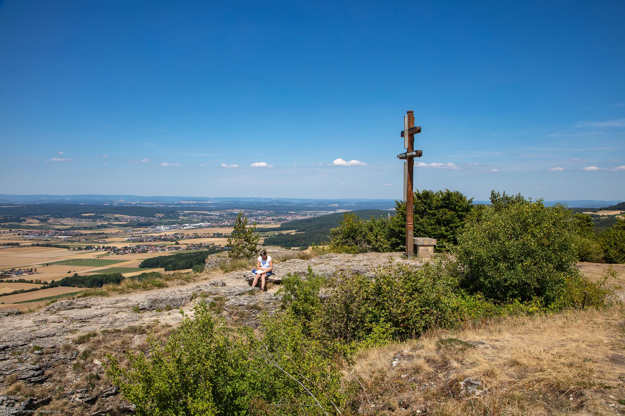 Frau sitzt auf Felsen neben großem Holzkreuz mit Blick auf Tal und Felder unter blauem Himmel.