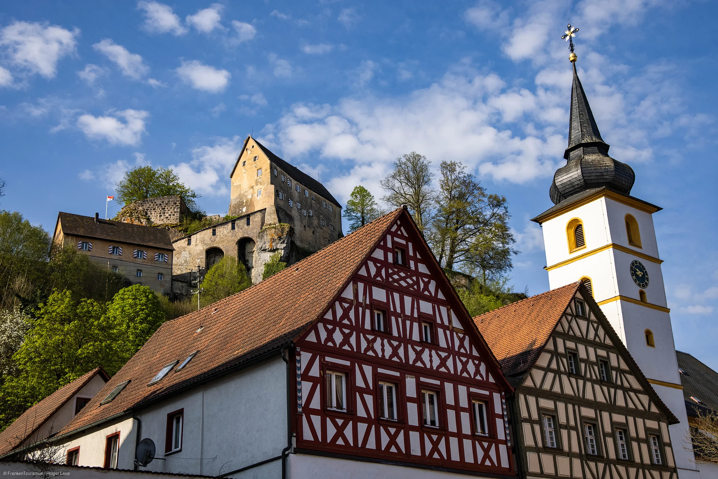 Fachwerkhäuser und Kirchturm vor bewaldetem Hügel mit Burg unter blauem Himmel und Wolken.