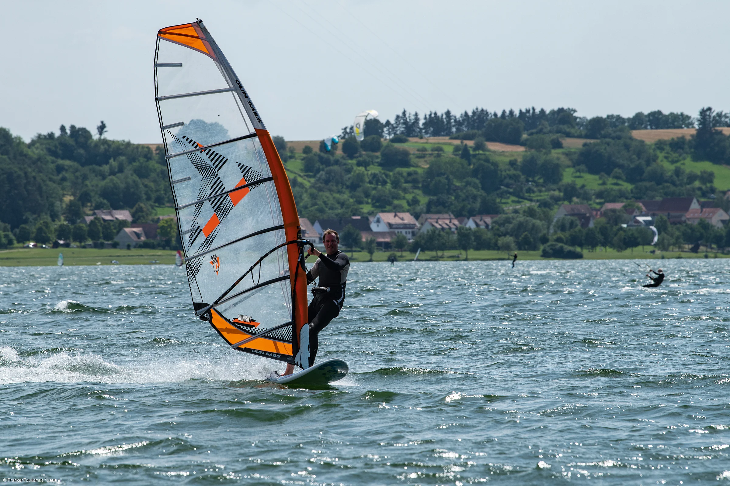 Mann beim Windsurfen auf einem See mit Ufer und Häusern im Hintergrund bei Tageslicht.