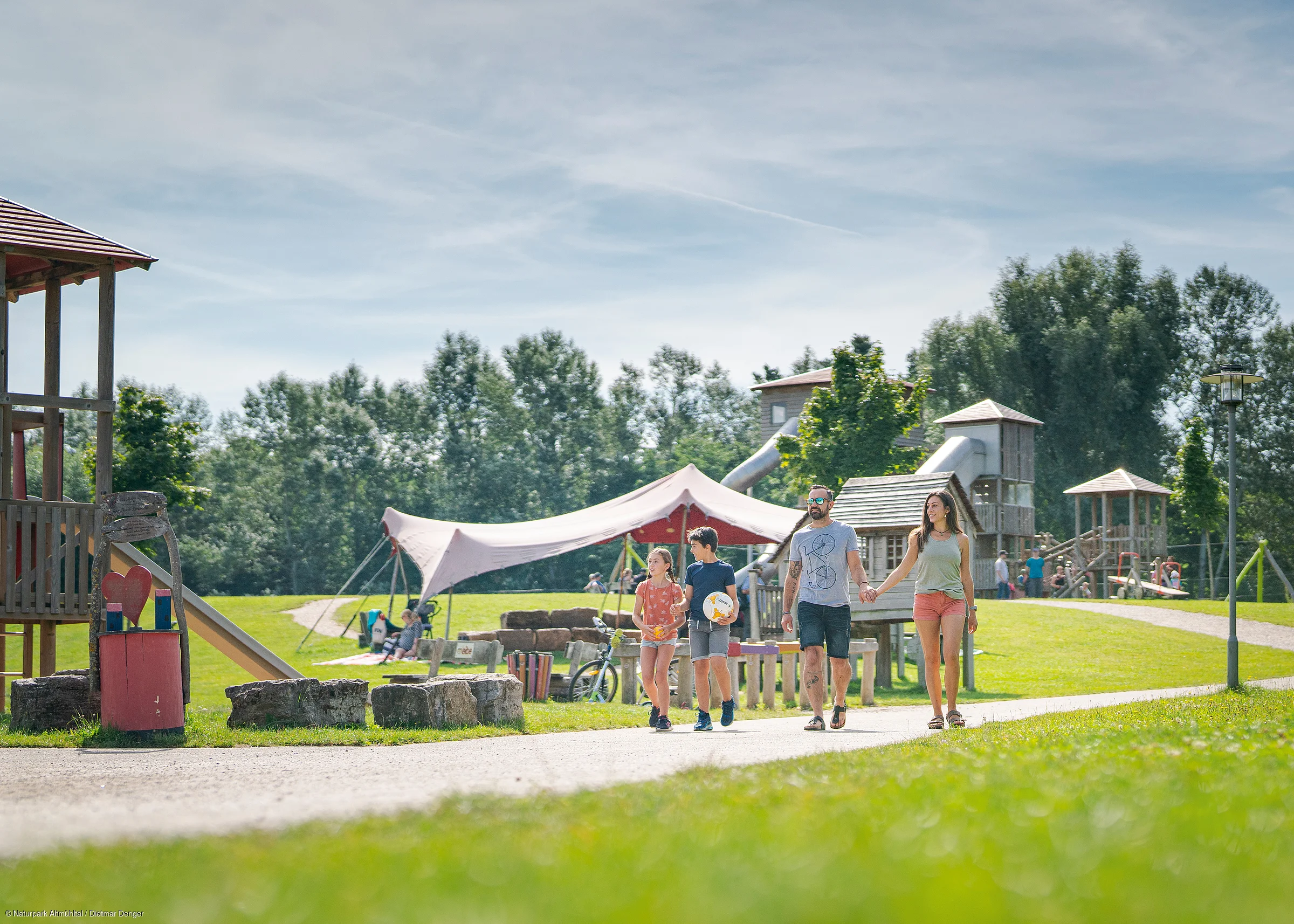 Familie mit zwei Kindern geht auf Weg in einem Park mit Spielplatz und Zelt bei sonnigem Wetter spazieren.