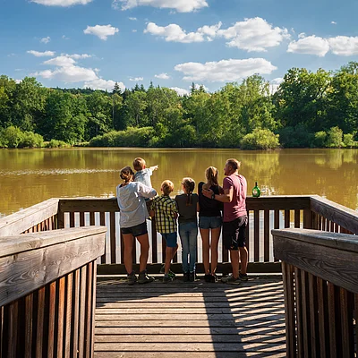 Sechs Personen stehen auf einem Holzsteg und blicken auf einen See mit bewaldetem Ufer bei sonnigem Himmel.